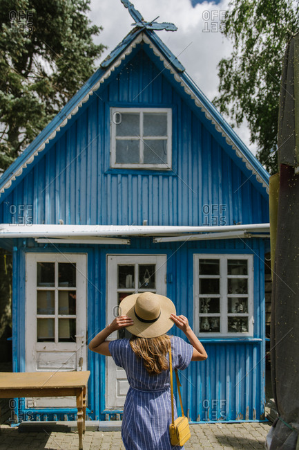 Back view of woman in straw hat and sundress standing in front of colorful blue summer house in daylight