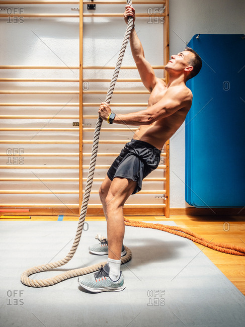 Side view of shirtless muscular sportsman climbing up rope while exercising in modern gym