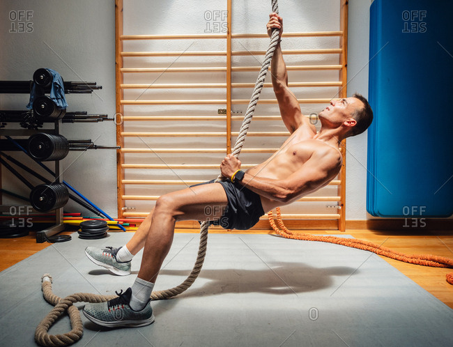 Side view of shirtless muscular sportsman climbing up rope while exercising in modern gym