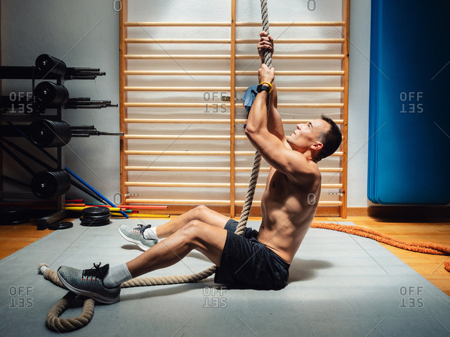 Side view of shirtless muscular sportsman climbing up rope while exercising in modern gym