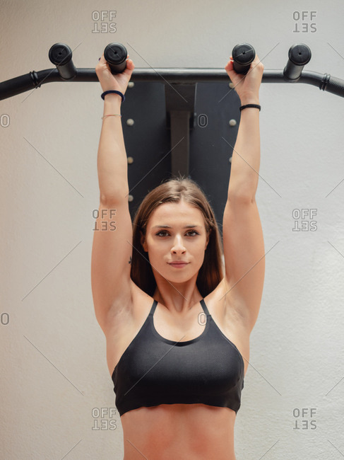 Muscular female athlete in sports bra looking at camera while hanging on bars during training in gym