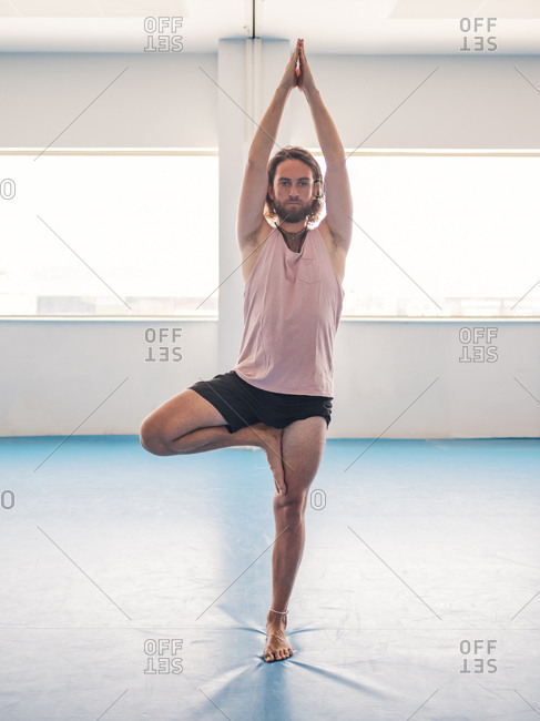 Bearded man in sportswear standing in one leg with prayer hands looking away on blue floor in studio
