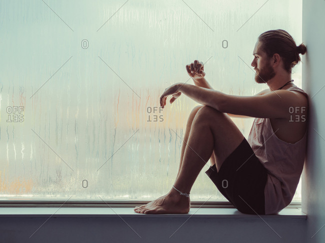 Side view of relaxed thoughtful male sitting in harmony on sill near against steamy window and looking down