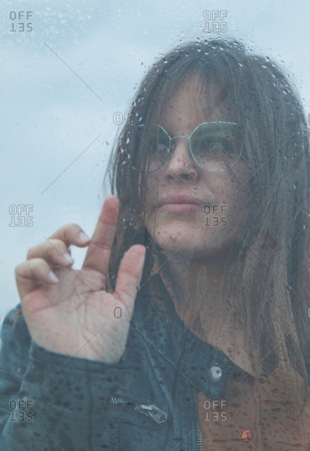 Portrait of beautiful woman with glasses looking out of wet window on rainy day looking away