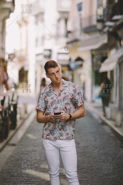Handsome happy male in hawaiian shirt standing on the street using mobile phone looking away