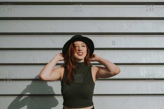 Attractive young woman with red hair in black hat holding hat and smiling on background of rough wall
