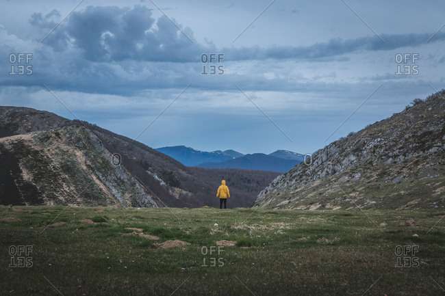 Back view of tourist in yellow coat looking at rough hills and overcast sky during trip in countryside
