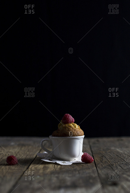 Ceramic cup with yummy raspberry muffin placed on shabby timber tabletop against black background