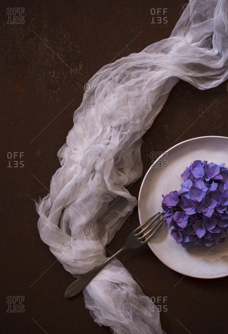 From above thin translucent fabric placed near plate with heap of violet flowers on brown surface