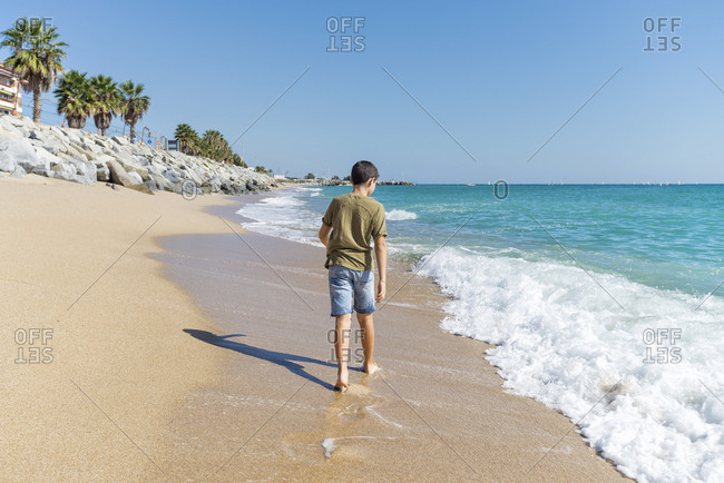 Rear view of young boy barefoot  walking on seashore in sunny day