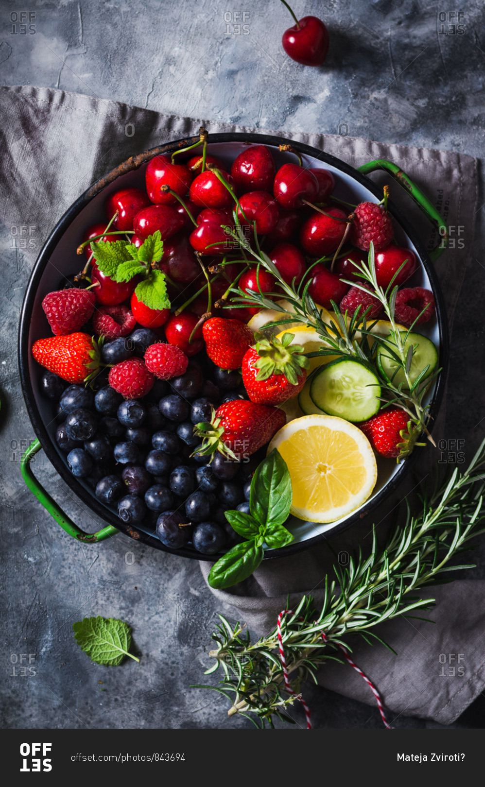 Variety of summer fruit and berries with fresh herbs, lemon and cucumber in rustic bowl on grey surface