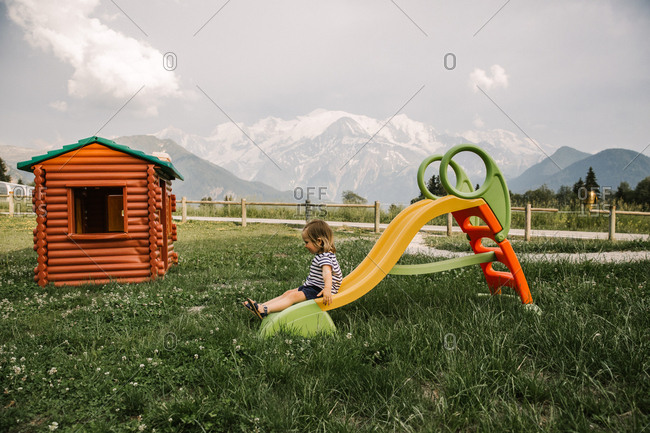 A toddler girl slides down a plastic slide in a field with a play house and snow capped mountains in the distance