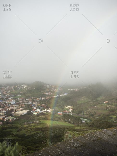 Rainbow against overcast foggy sky above town in green mountain valley