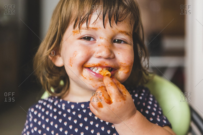 Close-up portrait of happy girl with messy face eating pasta while sitting at home