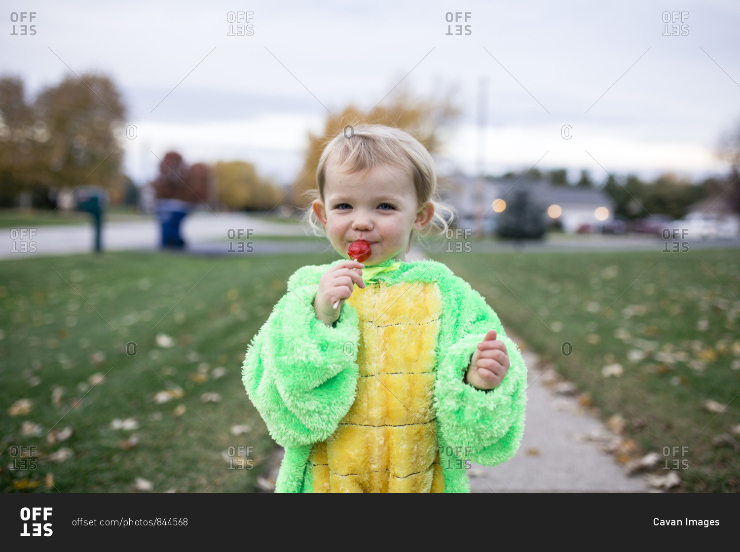 Happy toddler girl eating red sucker on sidewalk on Halloween