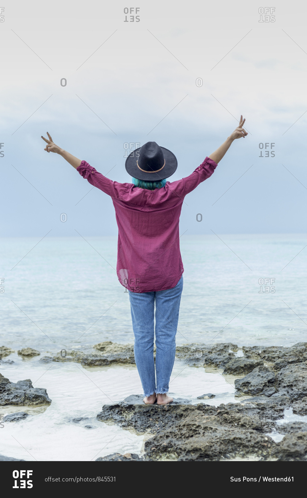 Back view of young woman with dyed blue hair standing on rock in front of the sea showing victory signs- Spain