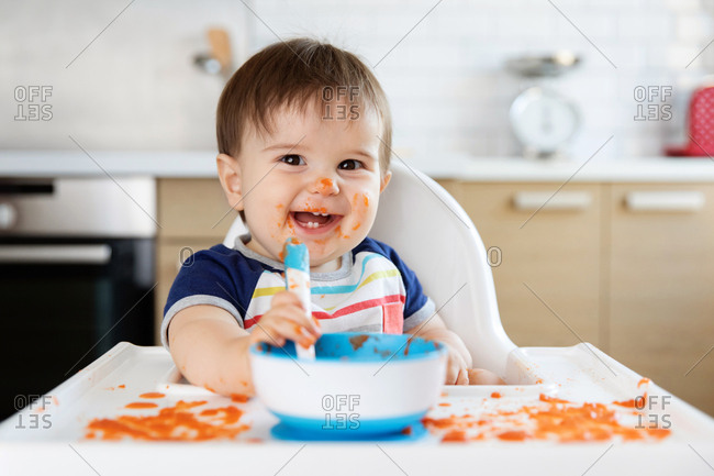 Baby boy in high chair eating pureed food with spoon - Stock Image