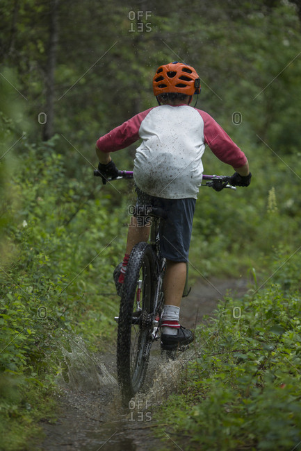 Boy, age 11 mountain biking through a puddle on a wet and muddy trail.