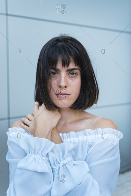 Woman in trendy blouse crossing hands and looking at camera against gray wall outside building