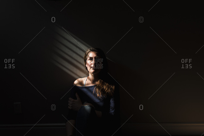 Portrait of woman sitting in a patch of striped light in a dark room.