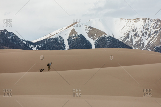 Man and dog walk over sand dunes with snowy mountains in background