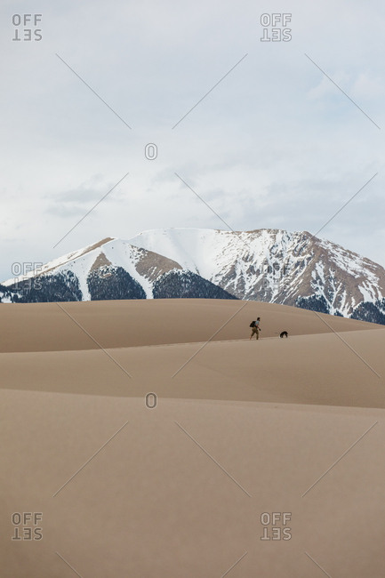 Male hiker with puppy hike the layered sand dunes under snowy mountain