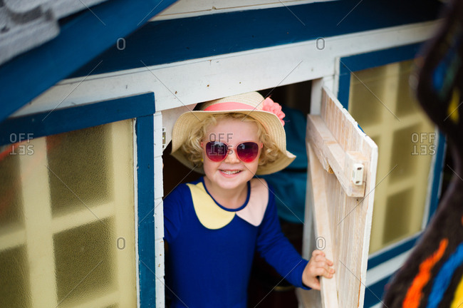 Amused little girl comes out of a little wooden house with a pink hat and glasses and a blue dress