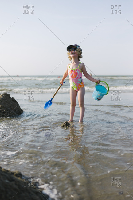 Little girl playing with a bucket and a shovel at the beach in the water and laughing loudly