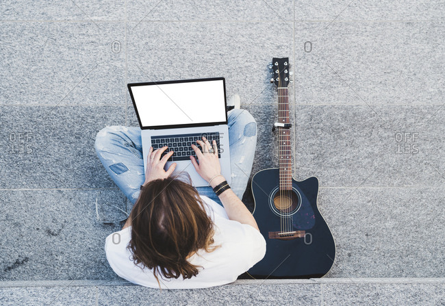 young man long hair writing on laptop sitting next acoustic guitar