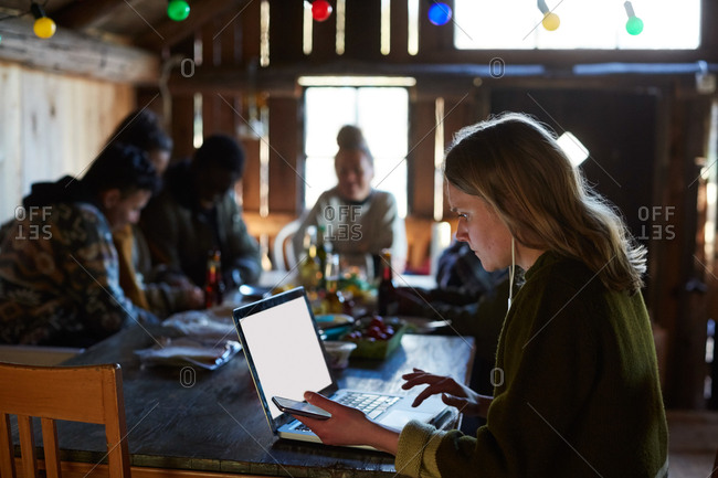 Young woman using laptop on table while friends talking in background at log cabin