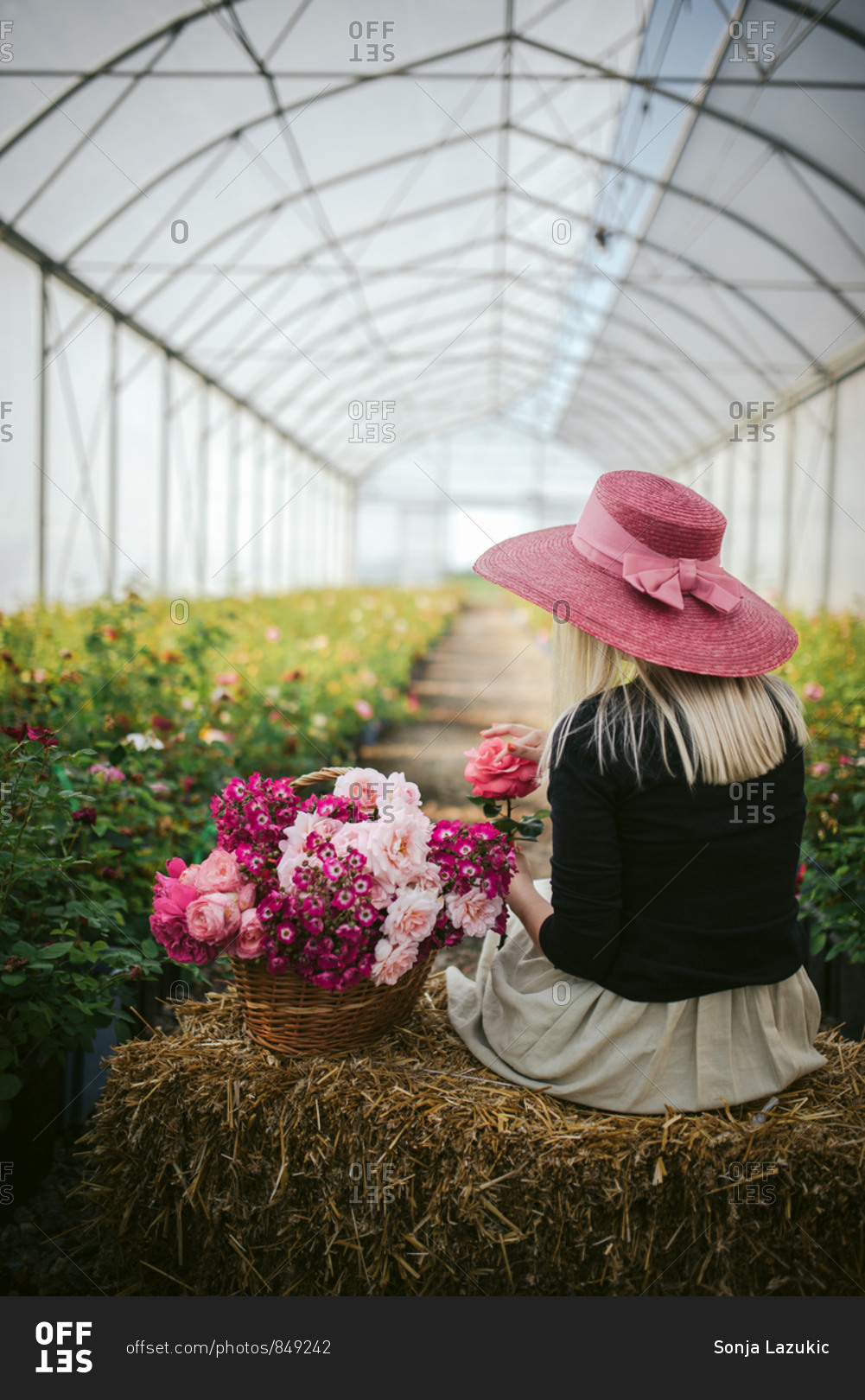 Woman in a pink hat by a basket of flowers on a bale of hay in a green house