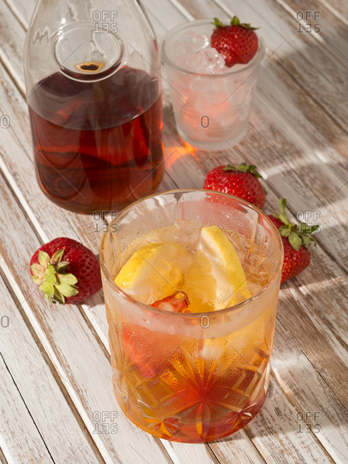 From above glass of iced drink with lemon and strawberry on rustic wooden table next to bottle in daylight