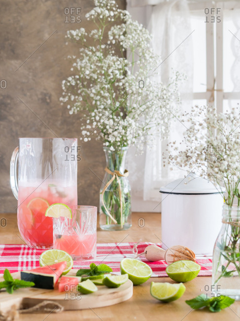 Cold glass jug of iced pink lemonade with watermelon and lime slices on rustic kitchen table next to bunch of gypsophila flowers in daylight