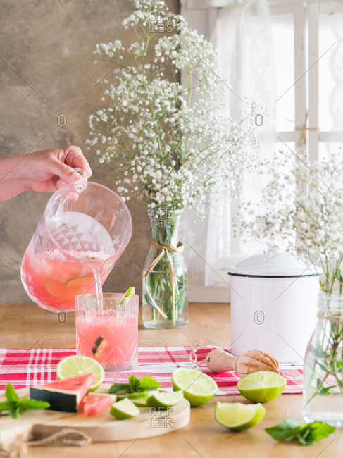 Iced pink drink with watermelon and lime slices pouring into glass from cold jar on rustic kitchen table next to bunch of gypsophila flowers in daylight
