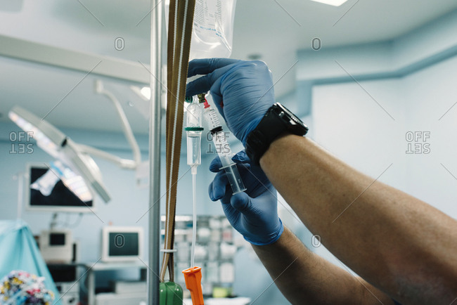 Crop hands in rubber gloves adding medication in saline solution drip with sterile syringe in hospital