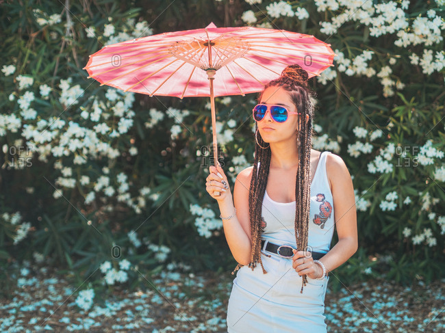 Beautiful slim young woman in summer outfit sunglasses and with umbrella standing legs crossed near blooming trees