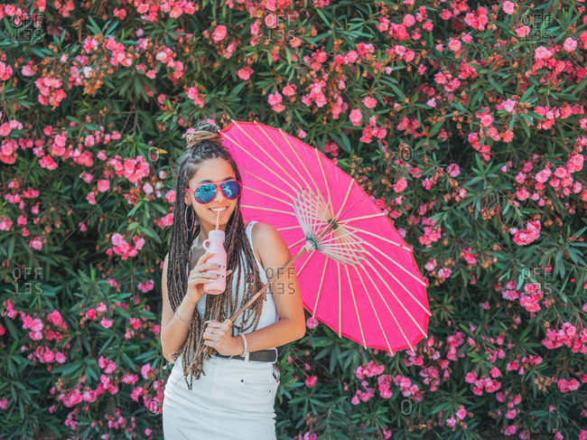 Beautiful slim young woman in summer outfit sunglasses and with umbrella drinking beverage near blooming trees