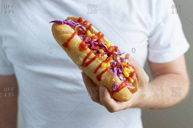 Male hand with a fresh hotdog topped with corn, red cabbage and chilies