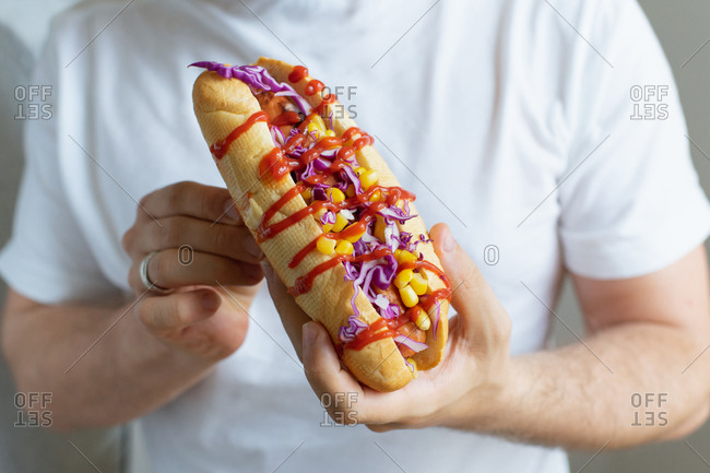 Male hand with a fresh hotdog topped with corn, red cabbage and chilies