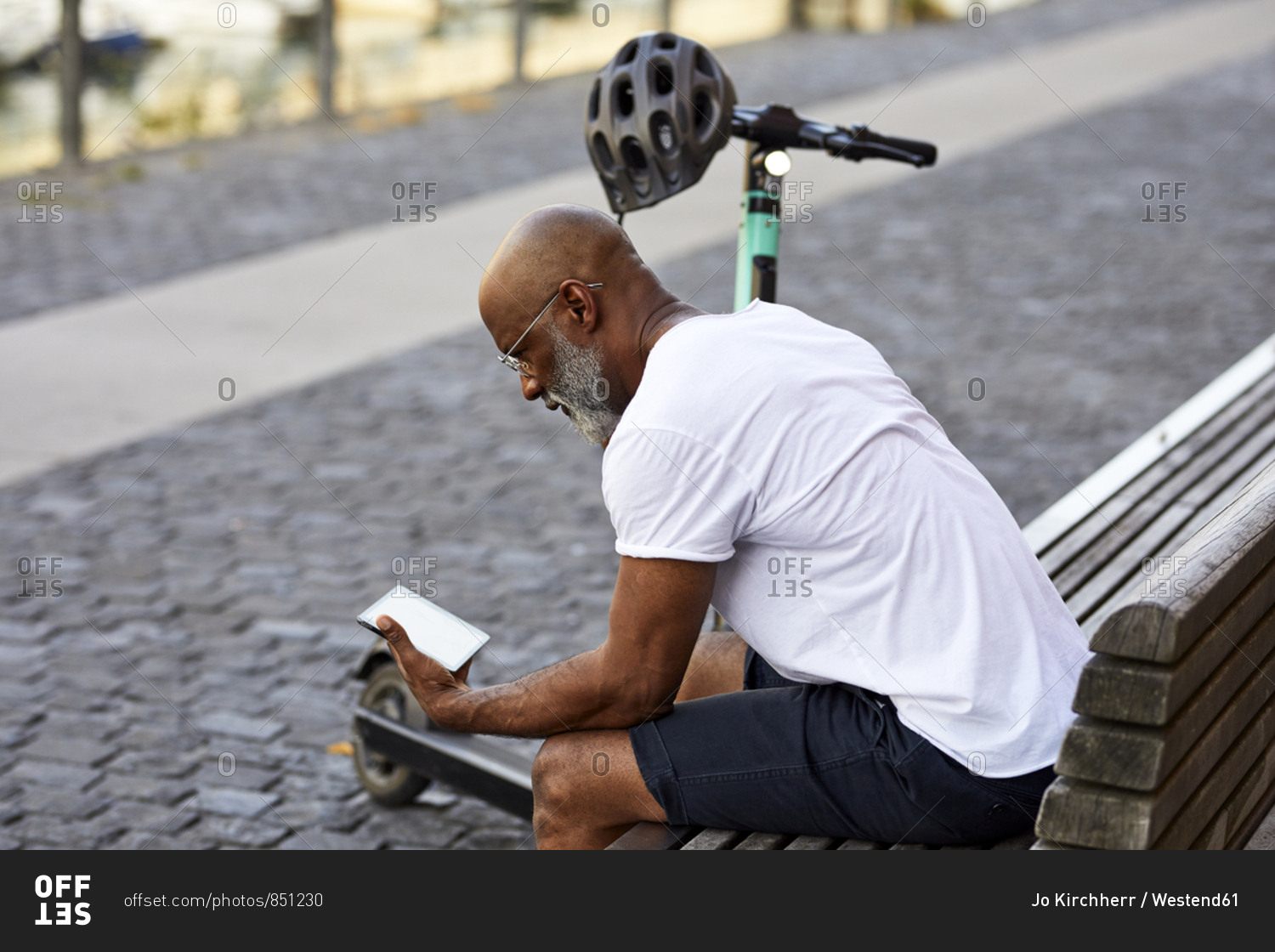 Mature man with E-Scooter sitting on bench in summer using mini tablet- Cologne- Germany