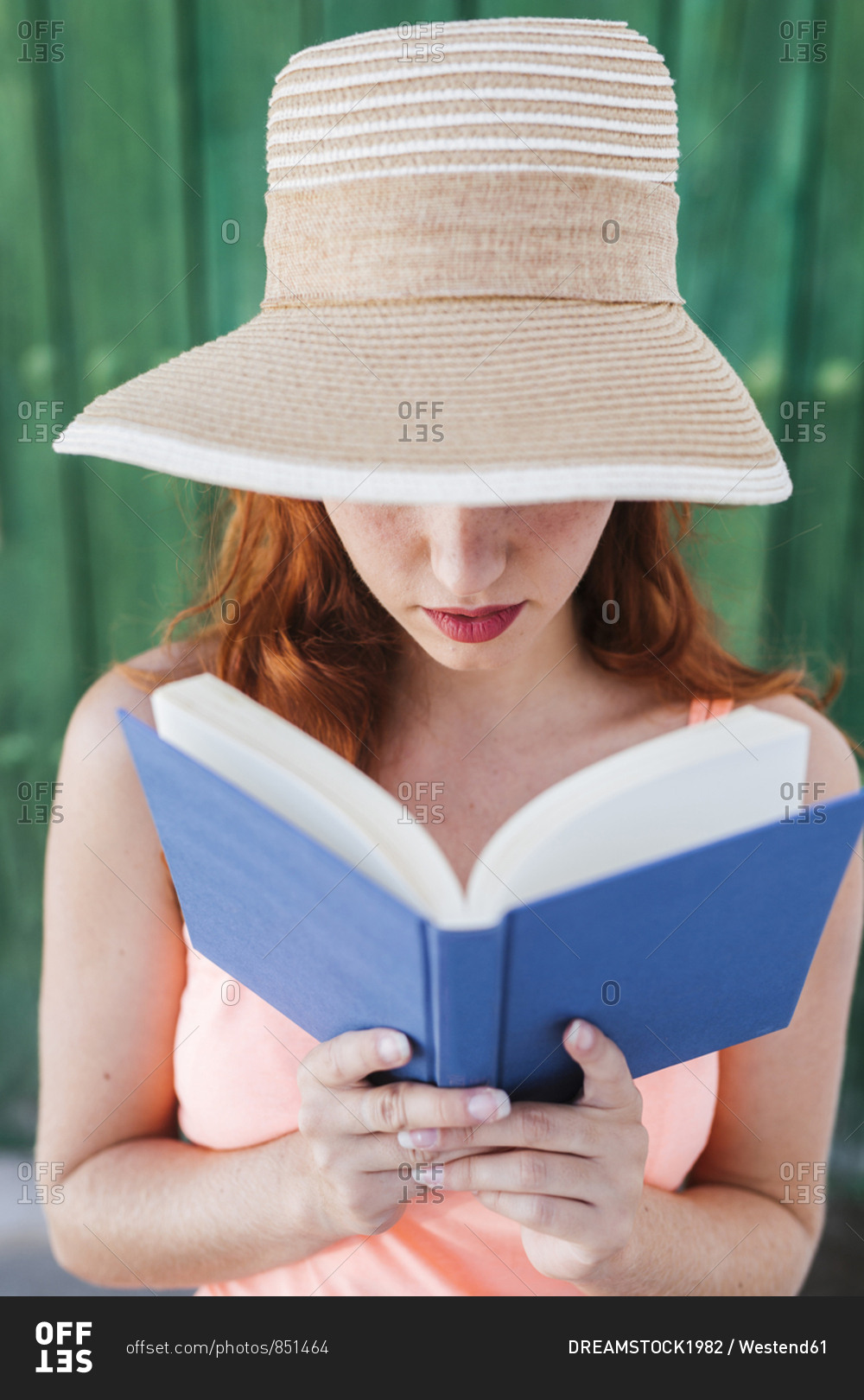 Redheaded young woman in front of green wooden door reading a book in summer