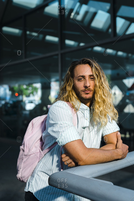 Happy long curly blonde haired guy in casual wear and pink backpack leaning on railing