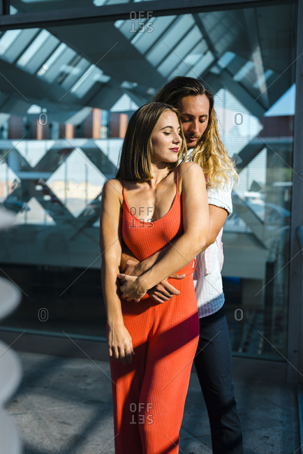 Happy long blonde haired guy embracing attractive young cheerful lady with red dress and looking down