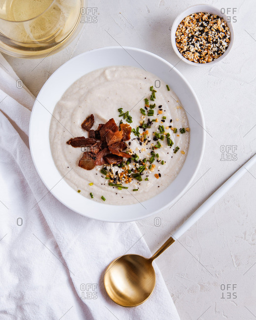 bowl of creamy winter soup garnished with chives and bacon, in a white bowl, on a white background, served with glass of white wine