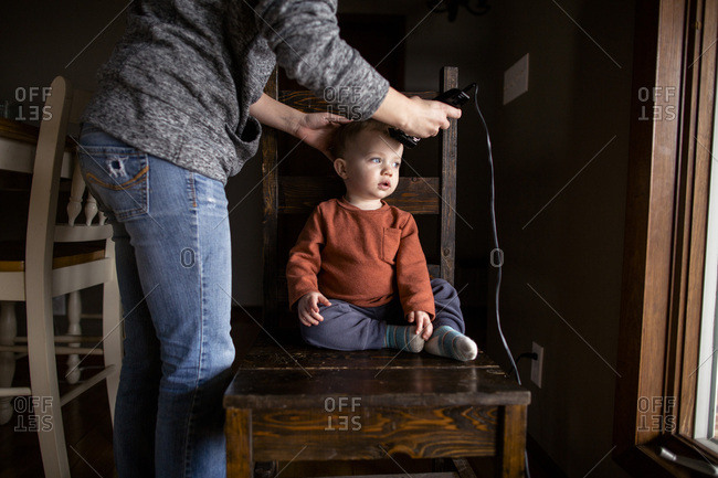 Mom cutting young boys hair inside sitting on chair next to window
