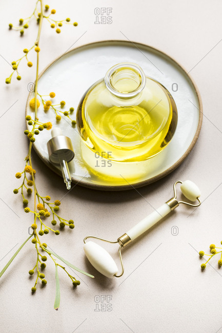 Bottle of oil on ceramic plate surrounded by yellow flowers and facial roller
