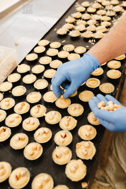 From above anonymous employee in glove putting fresh hazelnuts on top of yummy sweet pastry on tray while working in bakery
