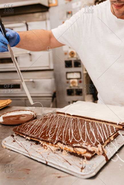 Crop man in latex glove and uniform decorating delicious cake with white cream swirls while working in bakery