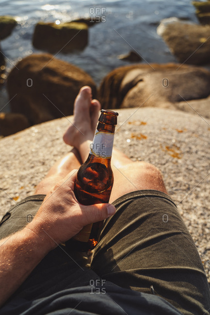 Crop man holding shiny bottle of beer resting sitting in stone on remote seashore