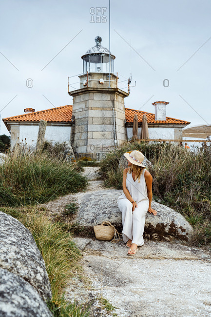 Traveling lady in white dress and hat standing alone on coastal stone in front of small lighthouse looking away